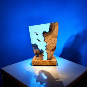 May include: A blue resin and wood sculpture of a diver in a blue ocean. The sculpture is on a wooden base and has a blue and white color scheme.