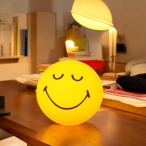 May include: A yellow smiley face night light with closed eyes and a wide smile. The light is on a wooden table.