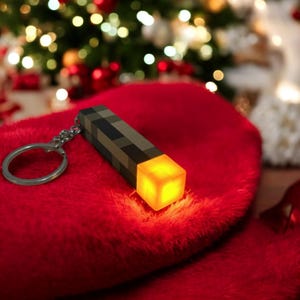 Op de afbeelding: Een gepixelde fakkel sleutelhanger met een gloeiende oranje kubus aan het einde. De sleutelhanger is zilverkleurig en de fakkel is bruin en zwart. De fakkel ligt op een rode, fuzzy ondergrond, met een kerstboom op de achtergrond.