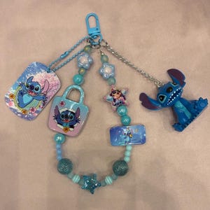 May include: A blue and pink keychain featuring the character Stitch from Lilo & Stitch. The keychain includes a dog tag, a purse charm, a star charm, and a rectangular charm, all with Stitch designs. A small Stitch figurine is also attached.