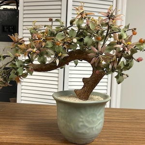 May include: A decorative bonsai tree sculpture featuring a textured brown trunk and branches adorned with carved green leaves, pale pink flowers, and small orange and white spherical accents, all set in a light green ceramic pot with sand.