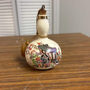 May include: A decorative, gourd-shaped bottle with a cork stopper and gold accents. The off-white ceramic bottle features a colourful illustration of a carriage and floral designs. A gold cord and tassel are attached to the bottle.