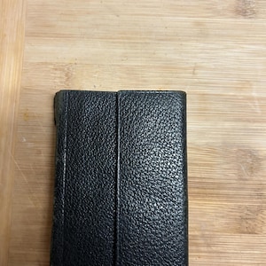 Puede incluir: Una cartera de cuero negro con una superficie texturizada. La cartera rectangular parece estar cerrada. El fondo es una superficie de madera de color marr&oacute;n claro.