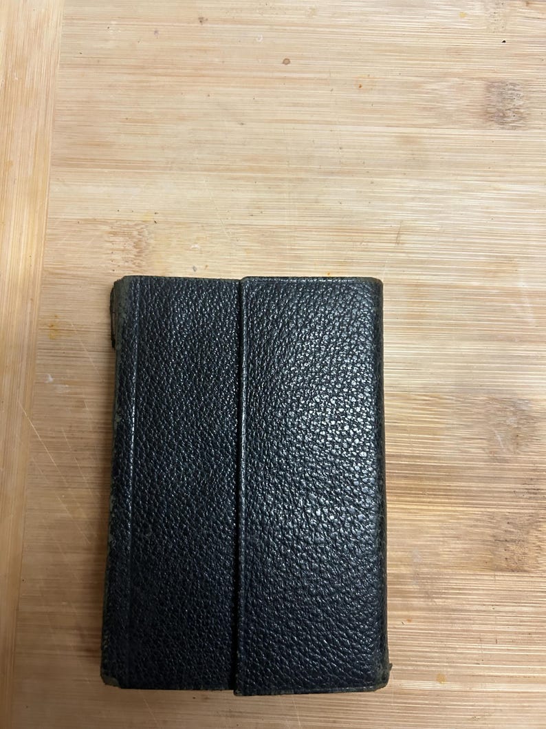 Puede incluir: Una cartera de cuero negro con una superficie texturizada. La cartera rectangular parece estar cerrada. El fondo es una superficie de madera de color marr&oacute;n claro.
