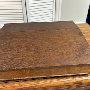 May include: A rectangular wooden box with a dark brown wood grain finish. The box features a hinged lid that spans the width of the top surface, with visible metal hinges along the back edge and a smooth, flat top panel construction.