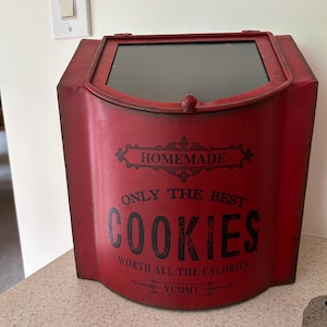 Peut inclure: Une boîte à biscuits en métal rouge avec un couvercle à charnière et une fenêtre transparente. L'avant présente un texte noir indiquant : HOMEMADE, ONLY THE BEST COOKIES, WORTH ALL THE CALORIES et YUMMY. Le récipient a une finition vieillie.
