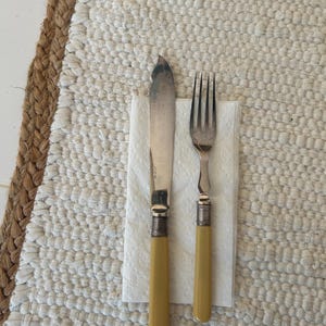 May include: A butter knife and fork set with yellow handles and silver blades and tines, resting on a white napkin. The silverware is placed on a textured, off-white rug with a brown border.