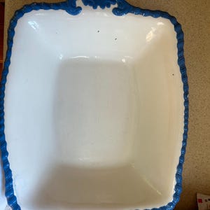 May include: A rectangular white ceramic baking dish featuring a raised, scalloped blue border with decorative scrollwork details along the edges. The interior of the dish is smooth and white, with a deep basin and a wide, textured blue rim.
