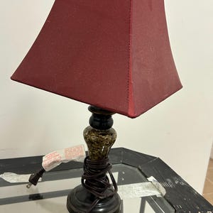 May include: A table lamp featuring a dark red, four-sided fabric shade and a decorative base with a mottled brown and black finish. The lamp includes a black power cord wrapped around the base and a white safety instruction tag attached to the cord.