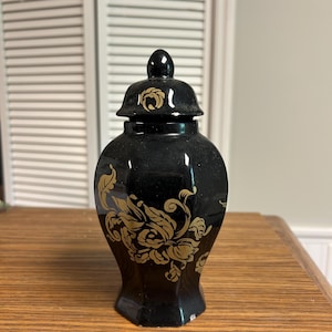 May include: A black ceramic ginger jar with a lid, featuring gold floral designs. The jar has a glossy finish and sits on a wooden surface. The jar's shape is slightly rounded with a hexagonal base.
