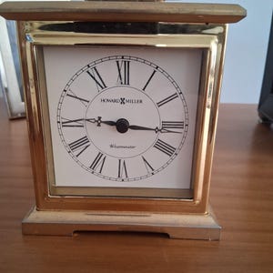 May include: A gold-toned rectangular clock with a white face and black Roman numerals. The clock face reads "HOWARD MILLER" and "Westminster". The clock has a handle on top and sits on a rectangular base.