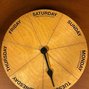 May include: A circular wooden day clock with a light brown grain finish, featuring black text for the days of the week from Monday to Sunday. A single black hand is centered on the face. Text includes WWW.DAYCLOCKS.COM and US PATENT #D433348.