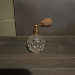 May include: A clear glass perfume bottle with a textured, honeycomb-like pattern on the body. The bottle features a gold-toned metal collar and a brown, fabric-covered bulb atomizer attached to the top, designed for dispensing fragrance.