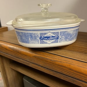 May include: An oval white ceramic casserole dish with a clear glass lid. The exterior features a blue marbled pattern, a blue horizontal stripe, and a central rectangular graphic with the text "The Art of Bon Appetit".