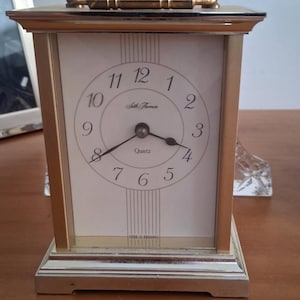 May include: A rectangular quartz clock with a gold-coloured frame and a white face. The clock has black numerals and hands, with the brand name "Seth Thomas". A gold handle is on top.
