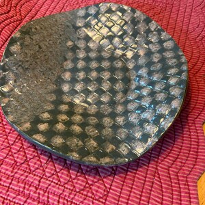 May include: A round ceramic plate featuring a dark green glaze with a textured, basket-weave pattern across the surface. The plate has a slightly irregular, organic edge and a glossy finish that highlights the raised grid design throughout the piece.