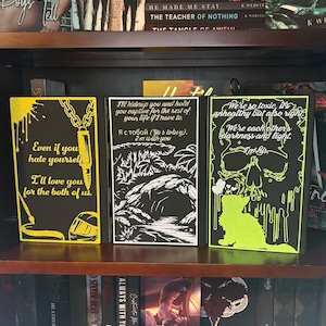 Puede incluir: Tres cubiertas de libros decorativas con fondos negros y acentos amarillos ne&oacute;n o verdes. Las cubiertas presentan citas e ilustraciones, incluyendo una calavera, un gato y una cadena. Los libros se exhiben en una estanter&iacute;a de madera con otros libros.
