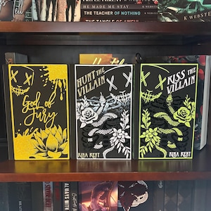 Puede incluir: Tres libros de tapa dura con cubiertas negras se exhiben en una estanter&iacute;a de madera. Los libros tienen dise&ntilde;os &uacute;nicos, incluyendo una flor de loto amarilla, una serpiente con detalles florales, y el texto "God of Fury", "Hunt the Villain" y "Kiss the Villain".
