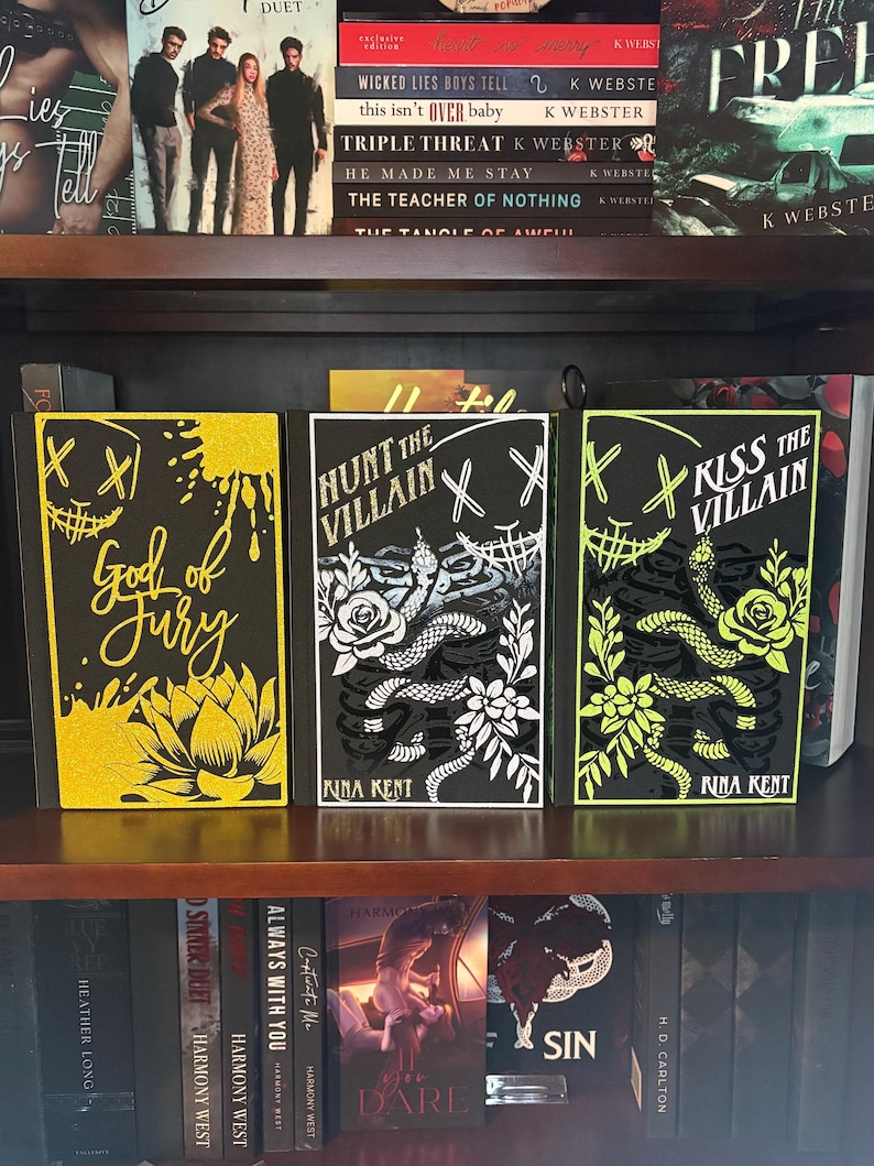 Puede incluir: Tres libros de tapa dura con cubiertas negras se exhiben en una estanter&iacute;a de madera. Los libros tienen dise&ntilde;os &uacute;nicos, incluyendo una flor de loto amarilla, una serpiente con detalles florales, y el texto "God of Fury", "Hunt the Villain" y "Kiss the Villain".