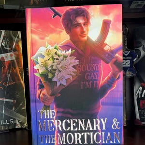 May include: Book cover for "The Mercenary & The Mortician" featuring a man holding a bouquet of white lilies and an assault rifle. The man wears a hoodie with the text "SOUNDS GAY, I'M IN." The title is in silver lettering.