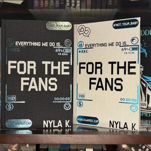 May include: Two hardcover books with the title "FOR THE FANS" in large white letters. One book is black, the other is off-white. Both books have the author's name, NYLA K., and other text and graphics. The books are on a wooden shelf.