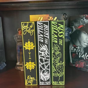 Puede incluir: Tres sujetalibros decorativos con los t&iacute;tulos "God of Fury", "Hunt the Villain" y "Kiss the Villain". Los sujetalibros son negros con detalles dorados, blancos y verde ne&oacute;n. El nombre de la autora, Rina Kent, tambi&eacute;n es visible.