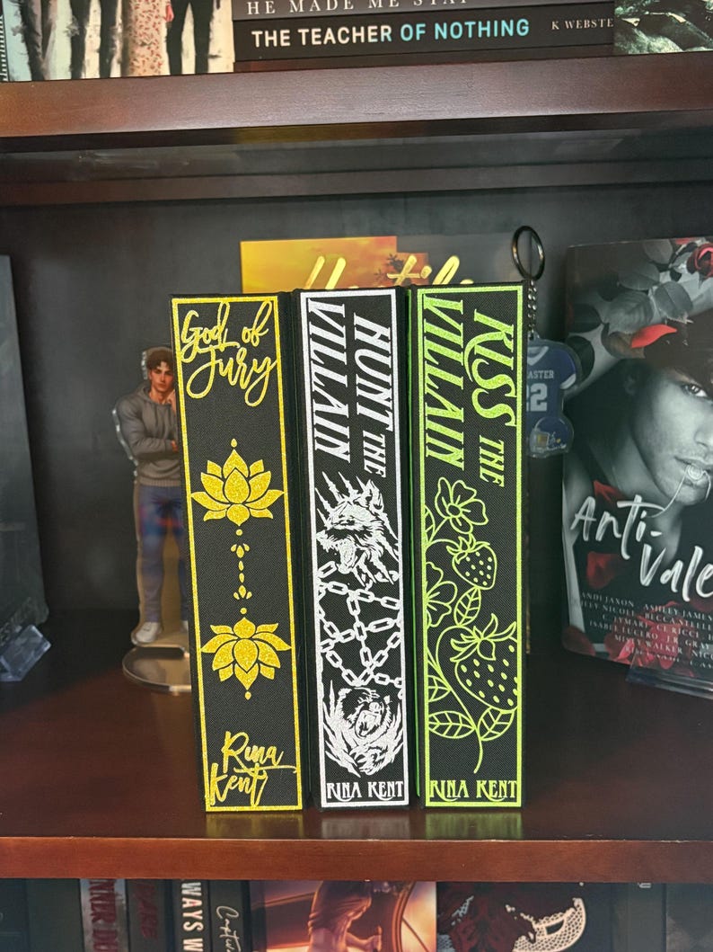 Puede incluir: Tres sujetalibros decorativos con los t&iacute;tulos "God of Fury", "Hunt the Villain" y "Kiss the Villain". Los sujetalibros son negros con detalles dorados, blancos y verde ne&oacute;n. El nombre de la autora, Rina Kent, tambi&eacute;n es visible.