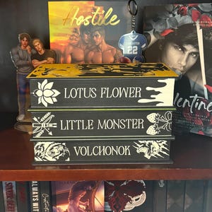 Puede incluir: Tres cajas decorativas negras con detalles dorados y blancos, cada una con un t&iacute;tulo: "LOTUS FLOWER", "LITTLE MONSTER" y "VOLCHONOK". Las cajas est&aacute;n en una estanter&iacute;a de madera, junto con otros libros y objetos decorativos.