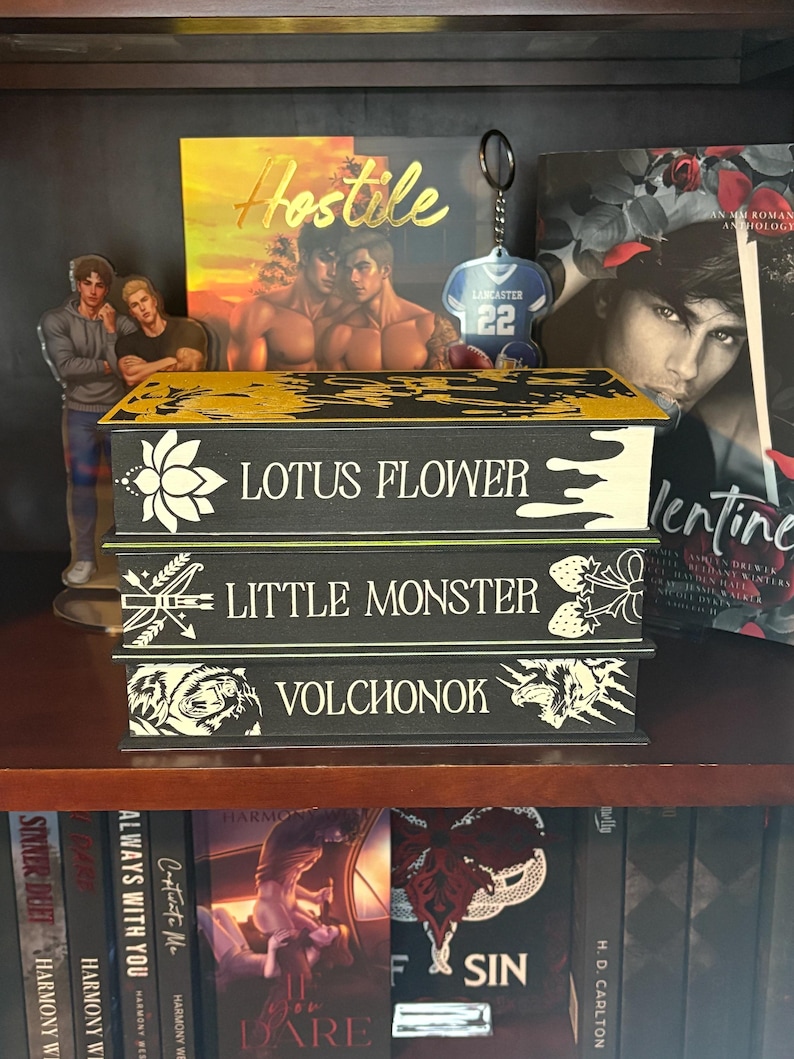 Puede incluir: Tres cajas decorativas negras con detalles dorados y blancos, cada una con un t&iacute;tulo: "LOTUS FLOWER", "LITTLE MONSTER" y "VOLCHONOK". Las cajas est&aacute;n en una estanter&iacute;a de madera, junto con otros libros y objetos decorativos.