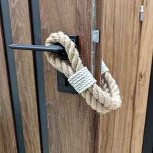 Op de afbeelding: Een houten deur met een zwarte deurknop en een touw eromheen gewikkeld. Het touw is in een knoop geknoopt en heeft een witte band eromheen.