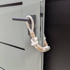 May include: A white rope doorstop with a knot tied in the middle. The rope is wrapped around a black door handle.
