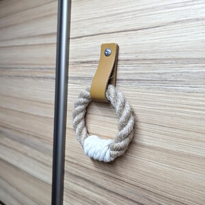 Op de afbeelding: Decoratieve kastgreep met een natuurlijke touwring met een witte accent, bevestigd aan een bruinleren band. De band is met een zilveren schroef aan een houten oppervlak bevestigd. De greep is voor een kast of lade.