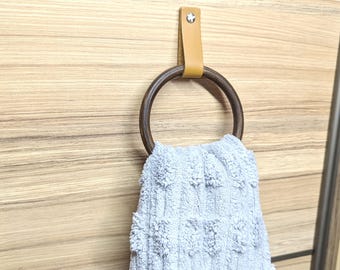 May include: A wooden towel ring with a brown leather strap hanging on a wood wall. A gray towel is hanging on the ring.