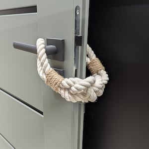 May include: A close-up of a door with a rope handle. The door is painted a light gray color and has a black door handle. The rope handle is made of white and brown rope and is wrapped around the door handle.