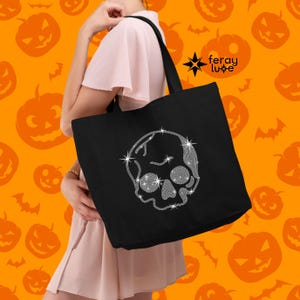 Puede incluir: Una bolsa de tela negra con un dise&ntilde;o de calavera plateada brillante. La bolsa tiene un asa larga y est&aacute; hecha de lona.