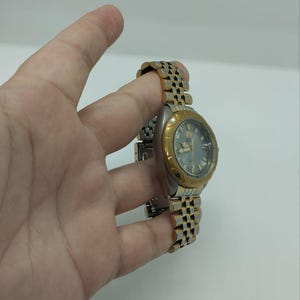 May include: A silver and gold-toned wristwatch with a silver dial. The watch has a gold-colored bezel and a two-tone metal band. The watch is held by a hand against a white background.