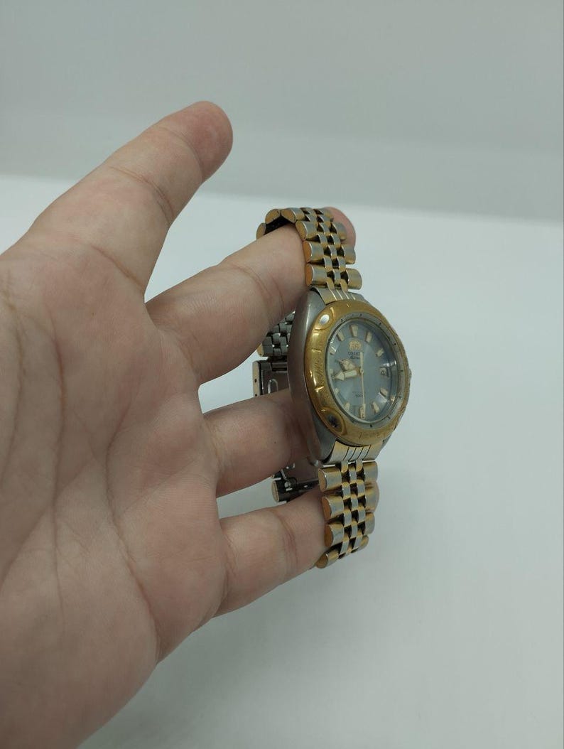 May include: A silver and gold-toned wristwatch with a silver dial. The watch has a gold-colored bezel and a two-tone metal band. The watch is held by a hand against a white background.