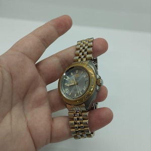 May include: A gold and silver-toned wristwatch with a silver dial. The watch has a gold bezel and a stainless steel band. The watch is held in a hand against a white background. The watch face displays the brand name "ORIENT".