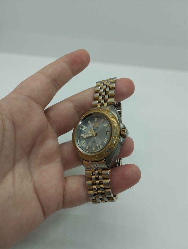 May include: A gold and silver-toned wristwatch with a silver dial. The watch has a gold bezel and a stainless steel band. The watch is held in a hand against a white background. The watch face displays the brand name "ORIENT".