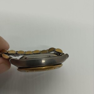 May include: A side view of a wristwatch with a silver-colored case and a gold-colored bezel. The watch band is a combination of silver and gold links. The watch is on a white background.