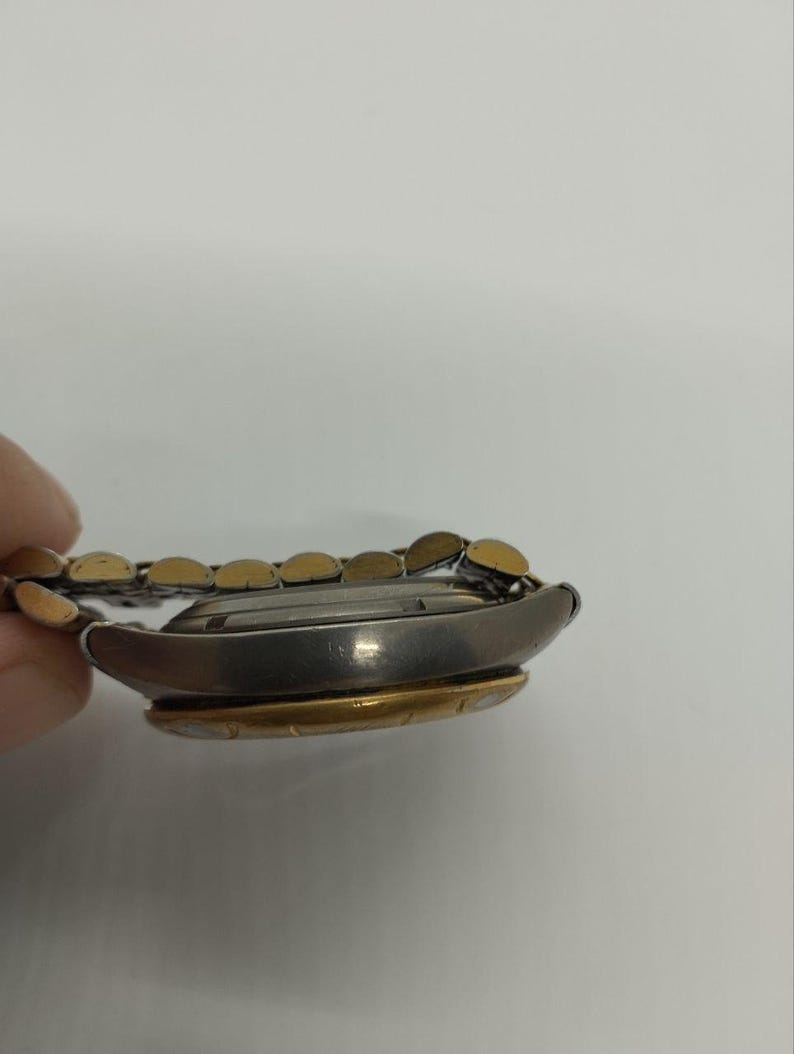 May include: A side view of a wristwatch with a silver-colored case and a gold-colored bezel. The watch band is a combination of silver and gold links. The watch is on a white background.
