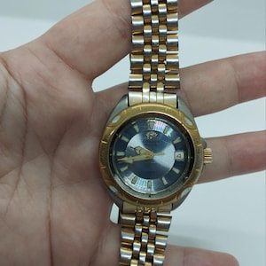 May include: A vintage Orient watch with a silver and gold-tone metal band and a blue face. The watch has a gold-tone bezel and a date display at the 3 o'clock position. The watch is held by a hand.