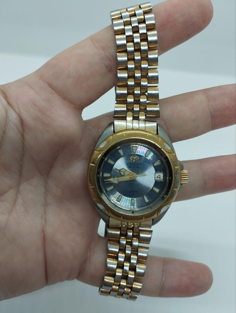 May include: A vintage Orient watch with a silver and gold-tone metal band and a blue face. The watch has a gold-tone bezel and a date display at the 3 o'clock position. The watch is held by a hand.