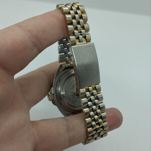 May include: A vintage wristwatch with a silver and gold-toned metal band. The watch band has a woven design. The watch face is visible, with a silver-toned case and a black dial. The watch is held by a hand.