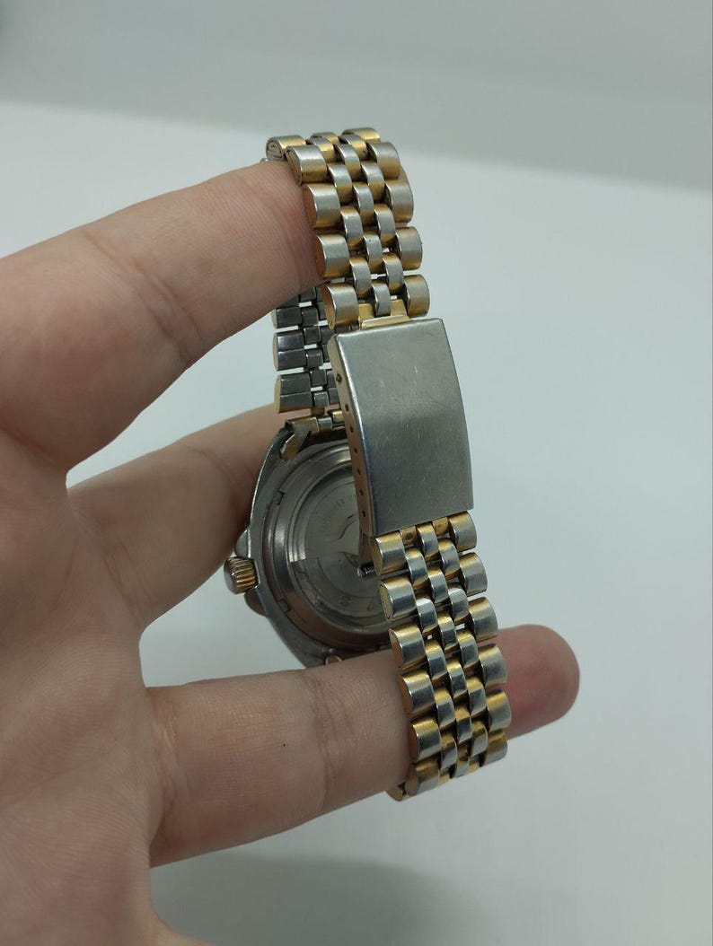 May include: A vintage wristwatch with a silver and gold-toned metal band. The watch band has a woven design. The watch face is visible, with a silver-toned case and a black dial. The watch is held by a hand.
