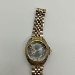 May include: A gold and silver-toned Orient watch with a gray face. The watch features a gold bezel and a silver and gold link band. The watch face displays the Orient logo, hands, and a date window. The watch is set against a white background.