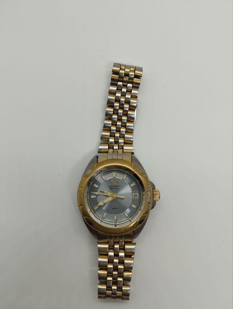 May include: A gold and silver-toned Orient watch with a gray face. The watch features a gold bezel and a silver and gold link band. The watch face displays the Orient logo, hands, and a date window. The watch is set against a white background.