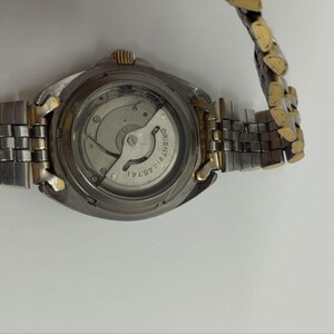 May include: A close-up of a wristwatch with a silver and gold-tone metal band. The watch face is visible, revealing the internal mechanisms. The watch has a round, silver-colored case and a gold-tone crown.