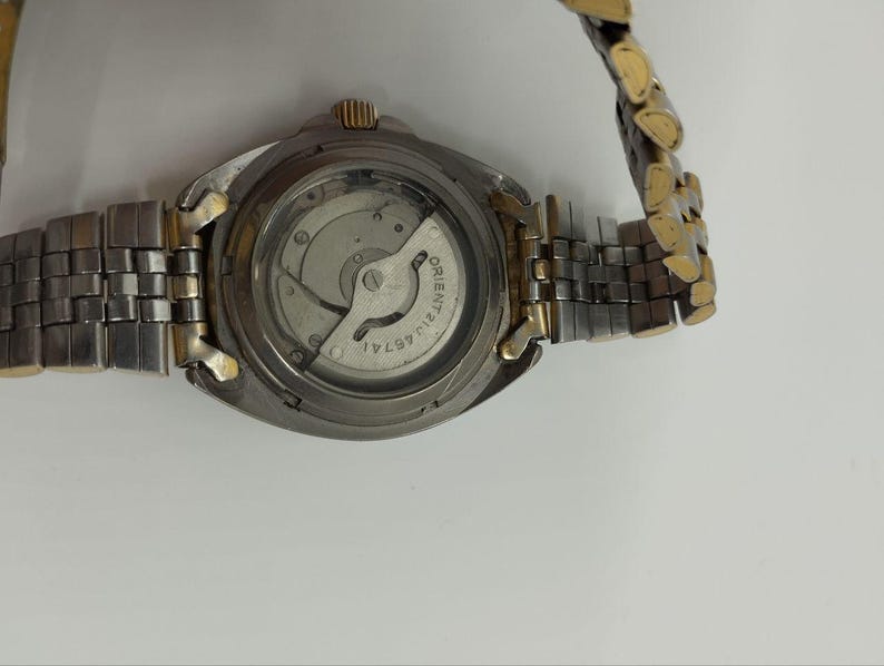 May include: A close-up of a wristwatch with a silver and gold-tone metal band. The watch face is visible, revealing the internal mechanisms. The watch has a round, silver-colored case and a gold-tone crown.