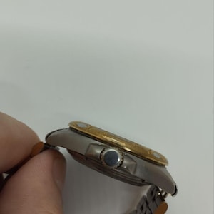 May include: A silver and gold wristwatch with a silver band. The watch face is silver with a gold bezel. The watch is being held by a person's hand.